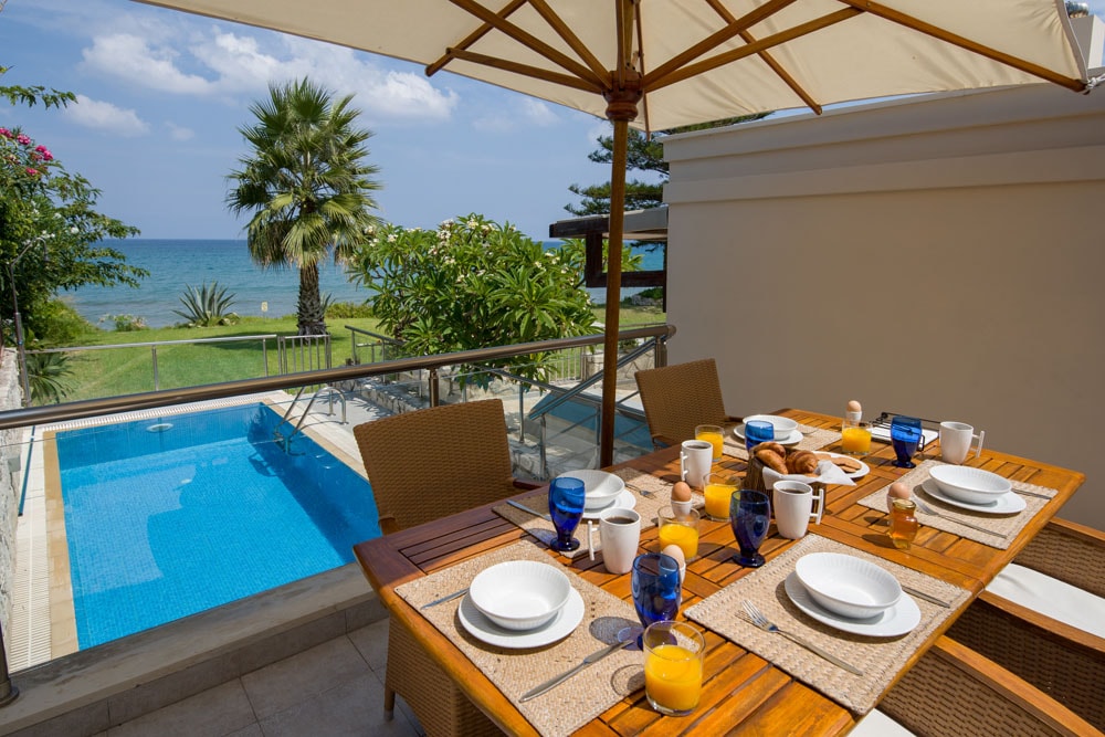 A dining table is set outdoors, featuring plates, glasses, and breakfast items. The table is positioned on a terrace overlooking a private pool and the ocean. Lush greenery and palm trees can be seen in the background, contributing to a refreshing coastal ambiance.