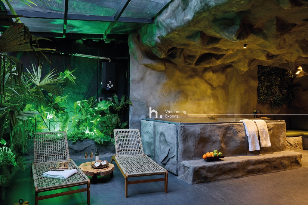 A cave-themed relaxation area is revealed, featuring a large jacuzzi surrounded by ambient green lighting. Two woven lounge chairs are positioned nearby, complemented by tropical plants that enhance the private atmosphere.