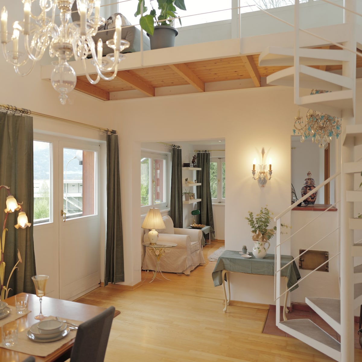 The bright interior features a wooden ceiling and large windows, allowing natural light to fill the space. A dining table is set for two, while a cozy seating area can be seen across the room. A spiral staircase leads to the upper level.