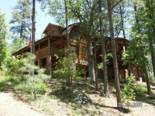 A large log and brick cabin is nestled among tall pine trees, showcasing a multi-level structure with a balcony. The natural setting provides a sense of seclusion, while the elevation of the property is apparent with sloping terrain and scattered foliage.