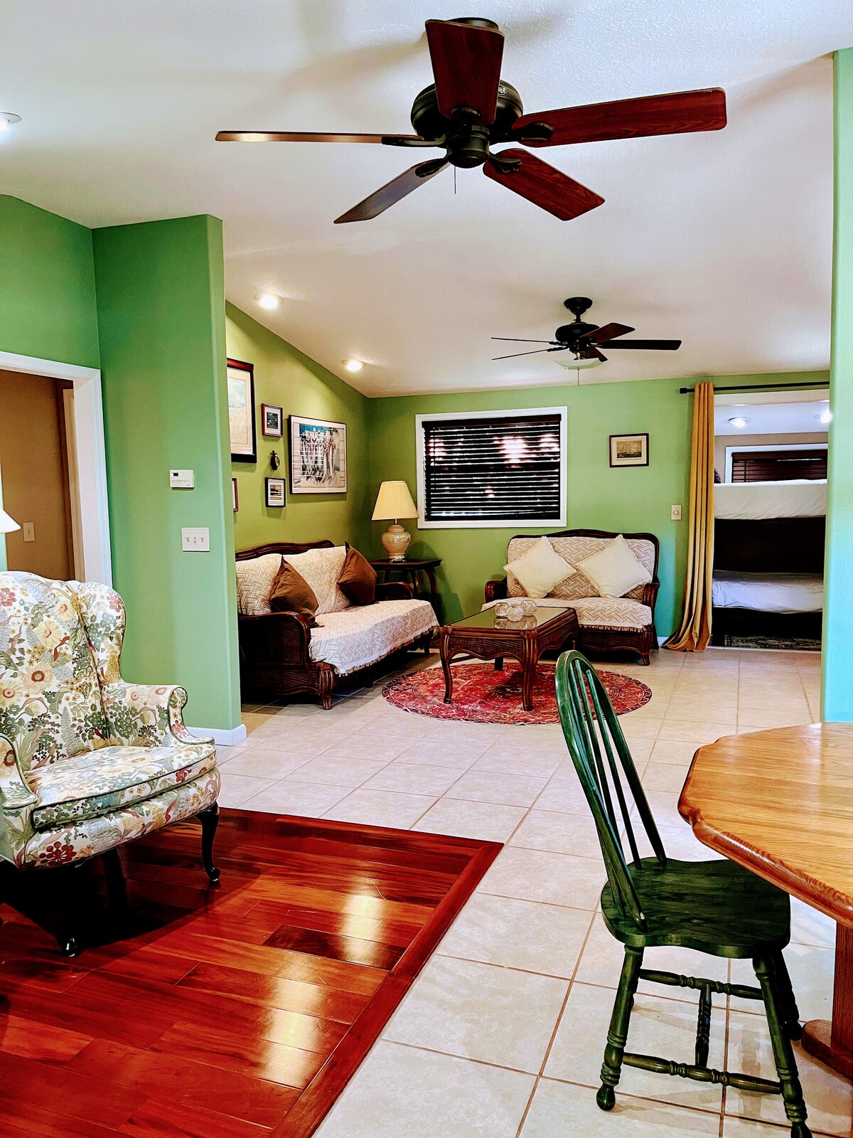 A seating area features two comfortable sofas and a pair of armchairs, arranged around a circular table on a patterned rug. Green walls and wooden accents create a welcoming environment. Ceiling fans circulate air while soft light is provided by nearby windows and lamps.