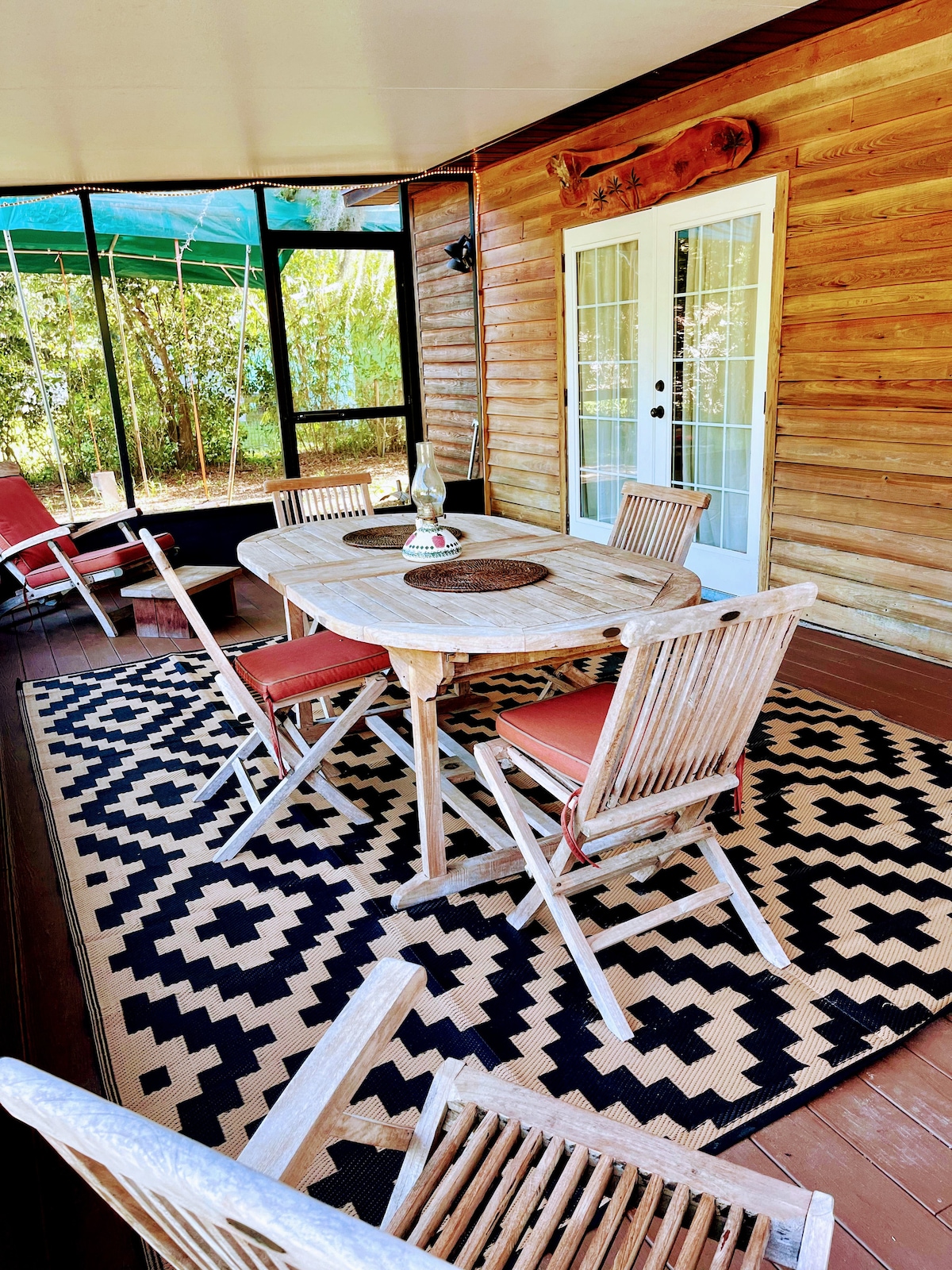 A screened porch features a rustic wooden table surrounded by six matching chairs. A woven rug with contrasting patterns lays beneath the table, while a decorative centerpiece rests in the middle. Light filters through the screens, revealing greenery outside.