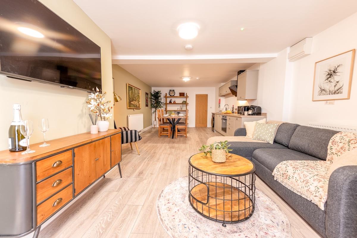 An open-plan living space features a comfortable lounge area with a sofa, side table, and a wall-mounted smart TV. A wooden dining table, accommodating up to four people, is visible beyond the kitchen, which includes modern appliances, all set against light-colored walls and wooden flooring.