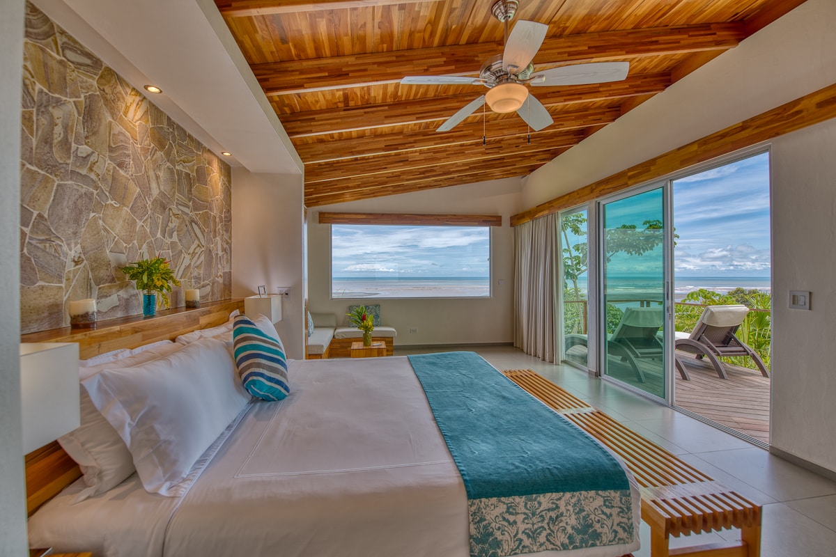 A spacious room features a large bed with a light blue throw and decorative pillows. Natural wood ceilings and a stone accent wall complement the panoramic windows showcasing ocean views. A private deck with seating is visible, enhancing the serene ambiance.