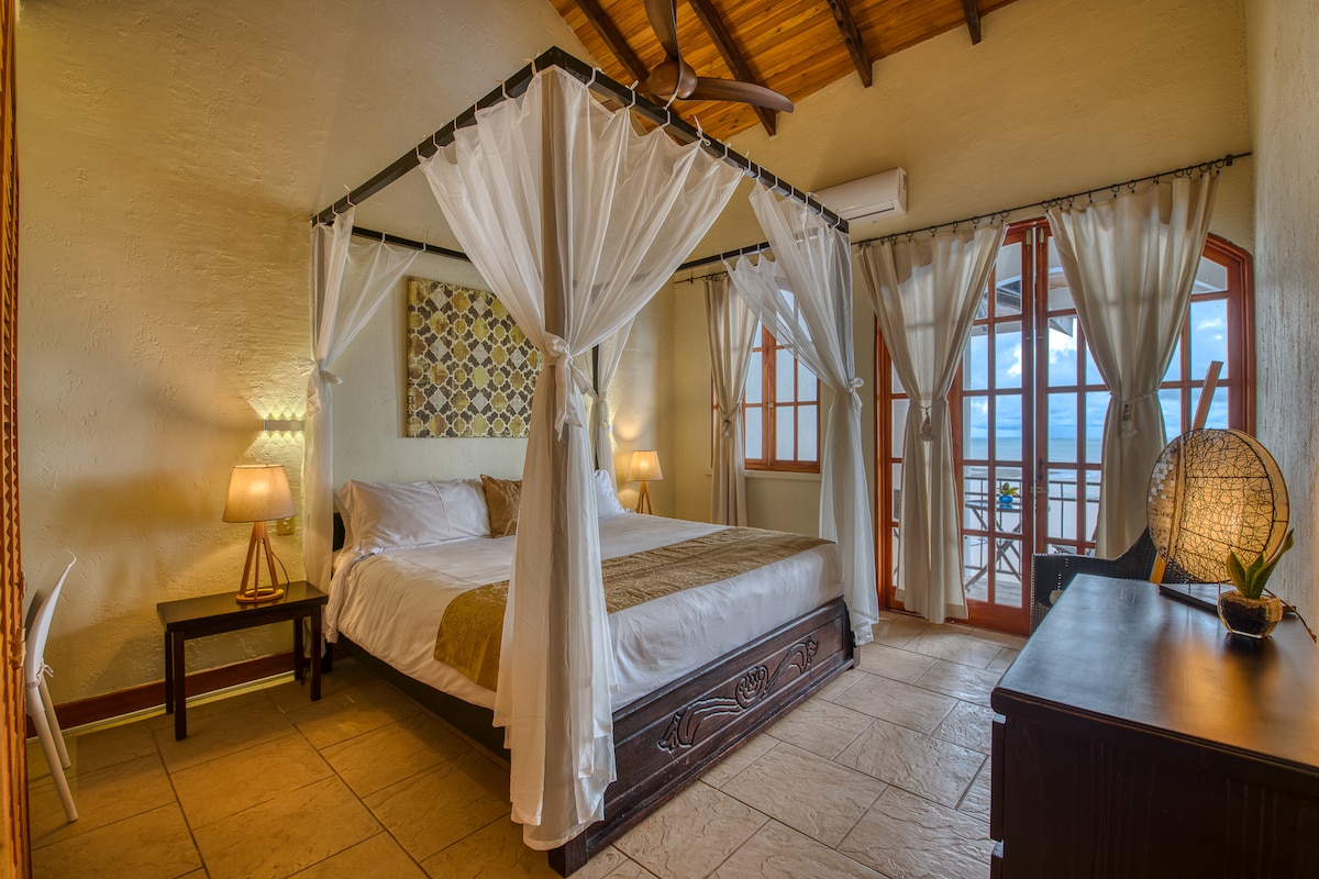 A spacious room features a king-size four-poster bed adorned with sheer drapery. Two bedside lamps provide soft illumination. French doors open to a balcony with ocean views, framed by wooden beams that add character to the high ceiling.
