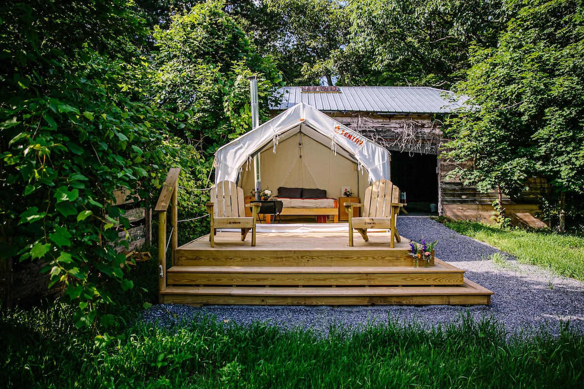 A spacious tent is situated on a wooden deck surrounded by lush greenery. Two Adirondack chairs are positioned in front of the tent, providing a cozy sitting area. The entrance is framed by a simple pathway leading up to the tent, enhancing the inviting outdoor space.