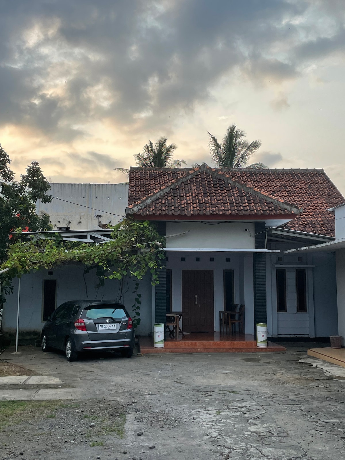A single-story house with a traditional tiled roof is visible, flanked by lush greenery. A vehicle is parked in front of the entrance, where two chairs are positioned near the door. A cloudy sky is seen above, hinting at the evening light.