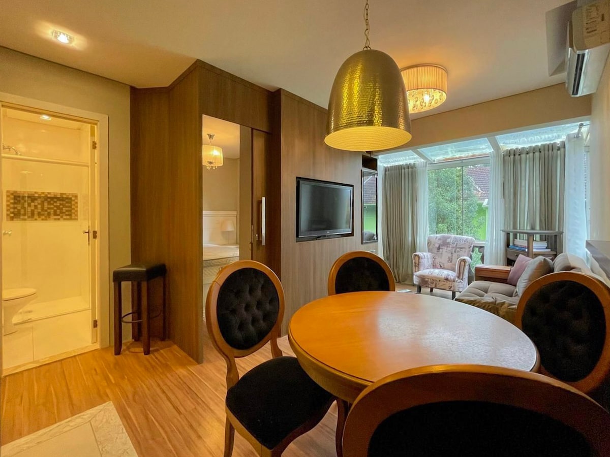 A living area is seen featuring a round dining table surrounded by four black chairs. A comfortable sofa is positioned nearby, with a large window providing natural light and views of the greenery outside. A mounted TV is visible on the wall.