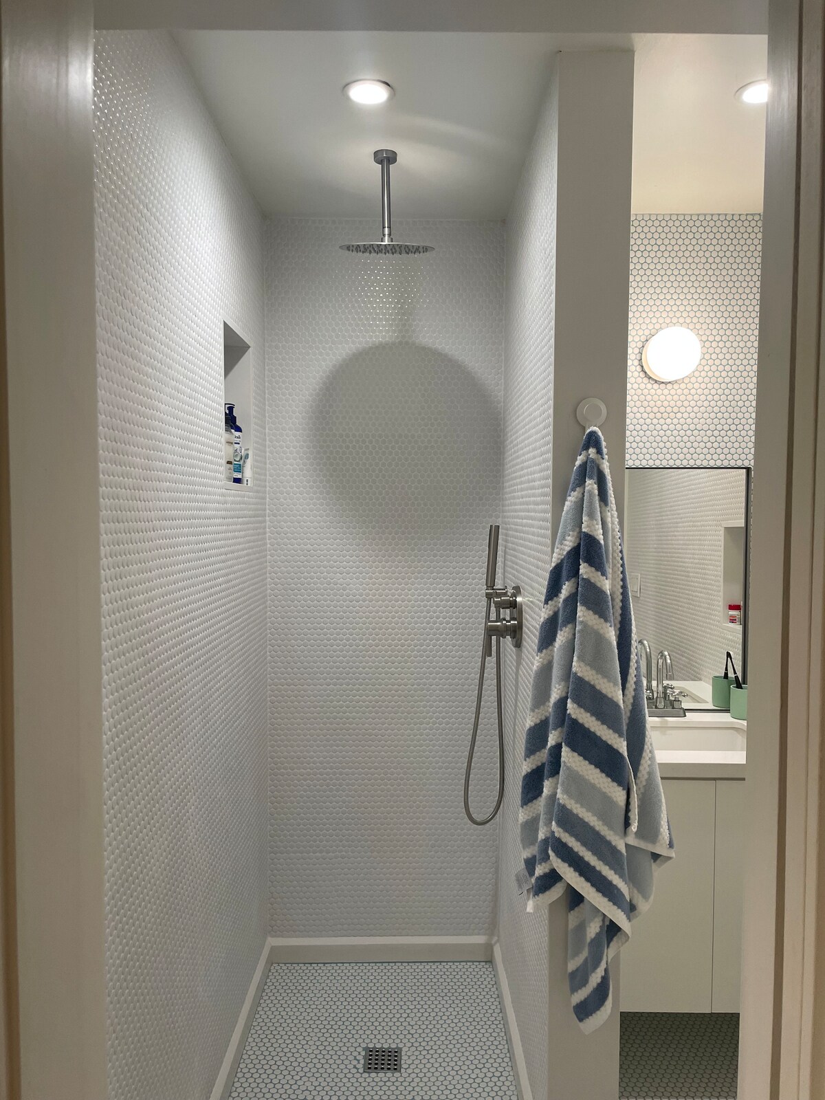 A modern shower area is enclosed by sleek white tile, featuring a wall-mounted showerhead. A striped towel is hung on a hook near the entrance, and a mirror reflects natural light. A minimalist vanity with storage is visible beside the shower.
