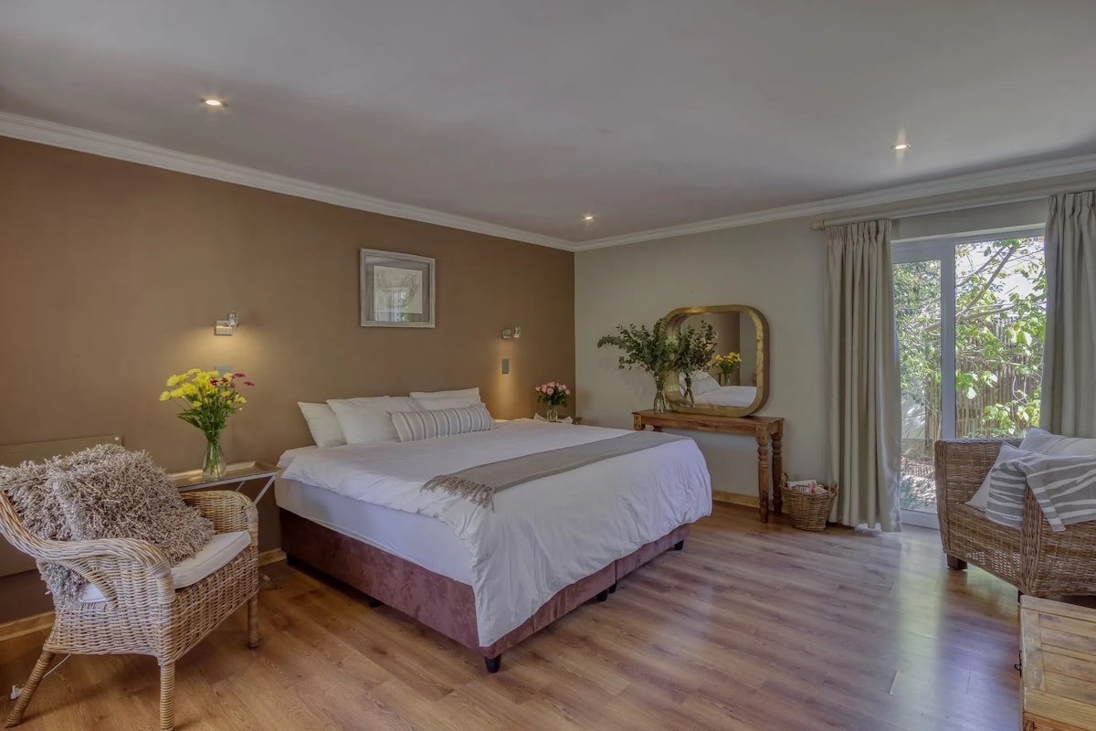 The spacious room features a large bed with white linens and a decorative throw. A full-length mirror reflects natural light from the sliding glass doors. Two wicker chairs with soft cushions and a wooden table enhance the inviting atmosphere, while plants add a touch of greenery.