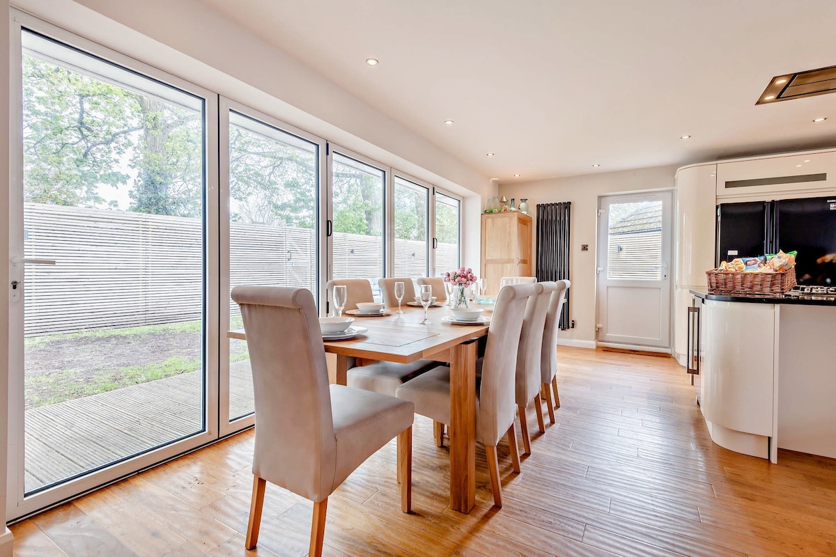 A spacious dining area is illuminated by natural light from large sliding doors that open to the garden. A wooden dining table is set for several diners, surrounded by cushioned chairs. An adjacent modern kitchen is visible, enhancing the open-plan layout.