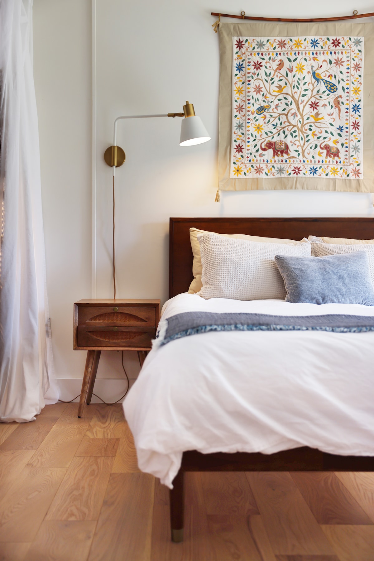 A cozy sleeping area features a wooden bed with soft linens and decorative pillows. A stylish lamp illuminates the space beside a small nightstand. A colorful wall hanging adds a vibrant touch above the bed, enhancing the room's character.