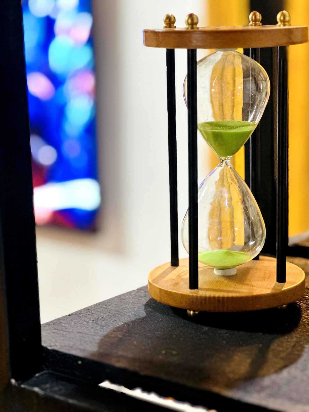 An elegant hourglass sits on a dark surface, featuring two glass chambers connected by a narrow neck. Soft green sand flows from the upper to the lower chamber, while a wooden base adds warmth to the design. A colorful, blurred background enhances the visual interest.