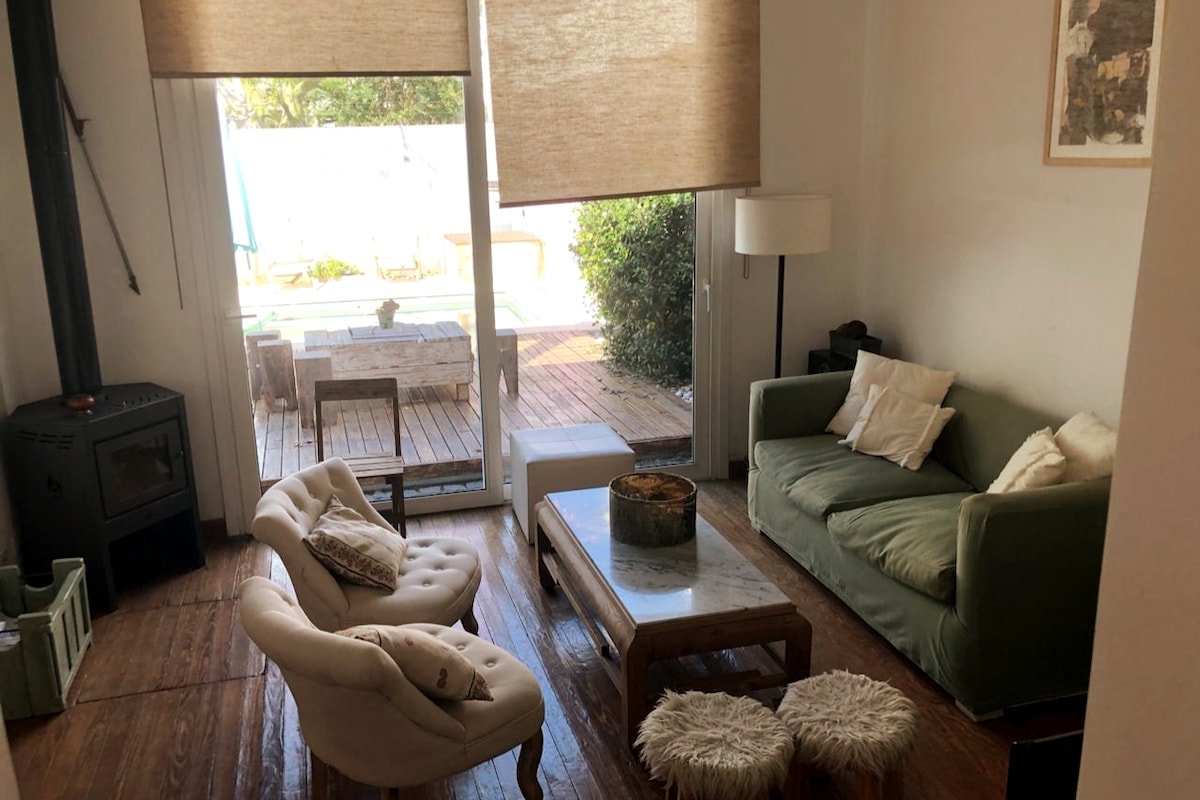 A comfortable living area features a green sofa and an elegant armchair positioned around a wooden coffee table. Natural light filters through large windows, illuminating the space. A cozy wood-burning stove adds warmth, while an outdoor deck is partially visible through the glass doors.
