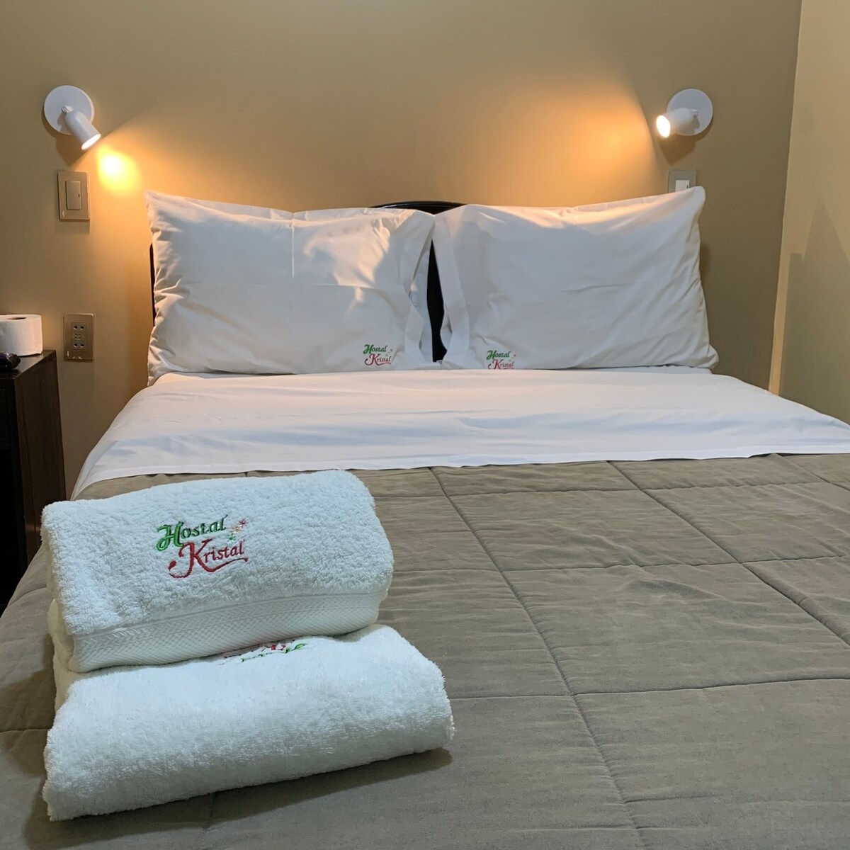 A neatly arranged bed is featured with plush white pillows against a soft beige wall. Two stylish wall lamps provide gentle illumination. Fresh towels are neatly stacked in the foreground, complementing the inviting atmosphere of the sleeping area.