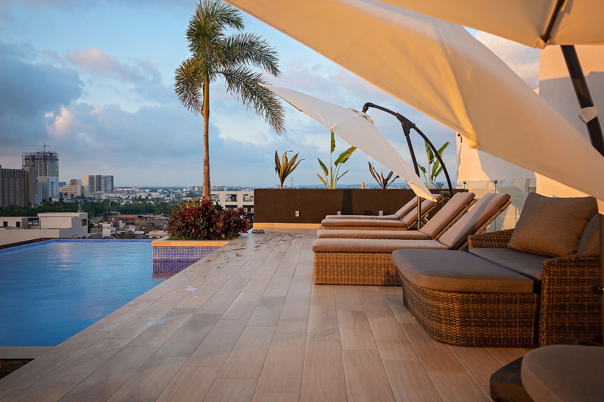 A rooftop area features an infinity pool with a stunning view of the city. Comfortable lounge chairs with cushions are arranged beside the pool. Large umbrellas provide shade, and tropical plants add greenery to the space, creating a serene environment for relaxation.