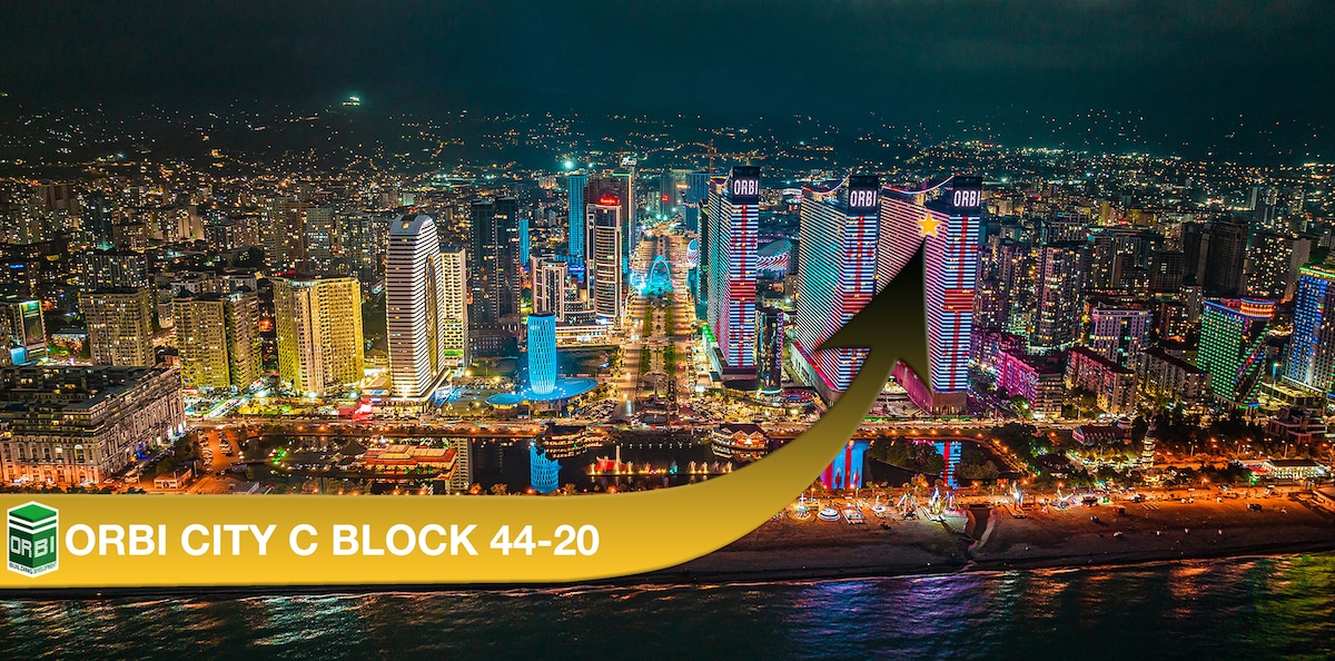 An aerial view of the vibrant Batumi coastline showcases the Orbi City complex illuminated at night. The buildings are adorned with colorful lights, creating a lively atmosphere, while the sandy beach and ocean are visible in the foreground.