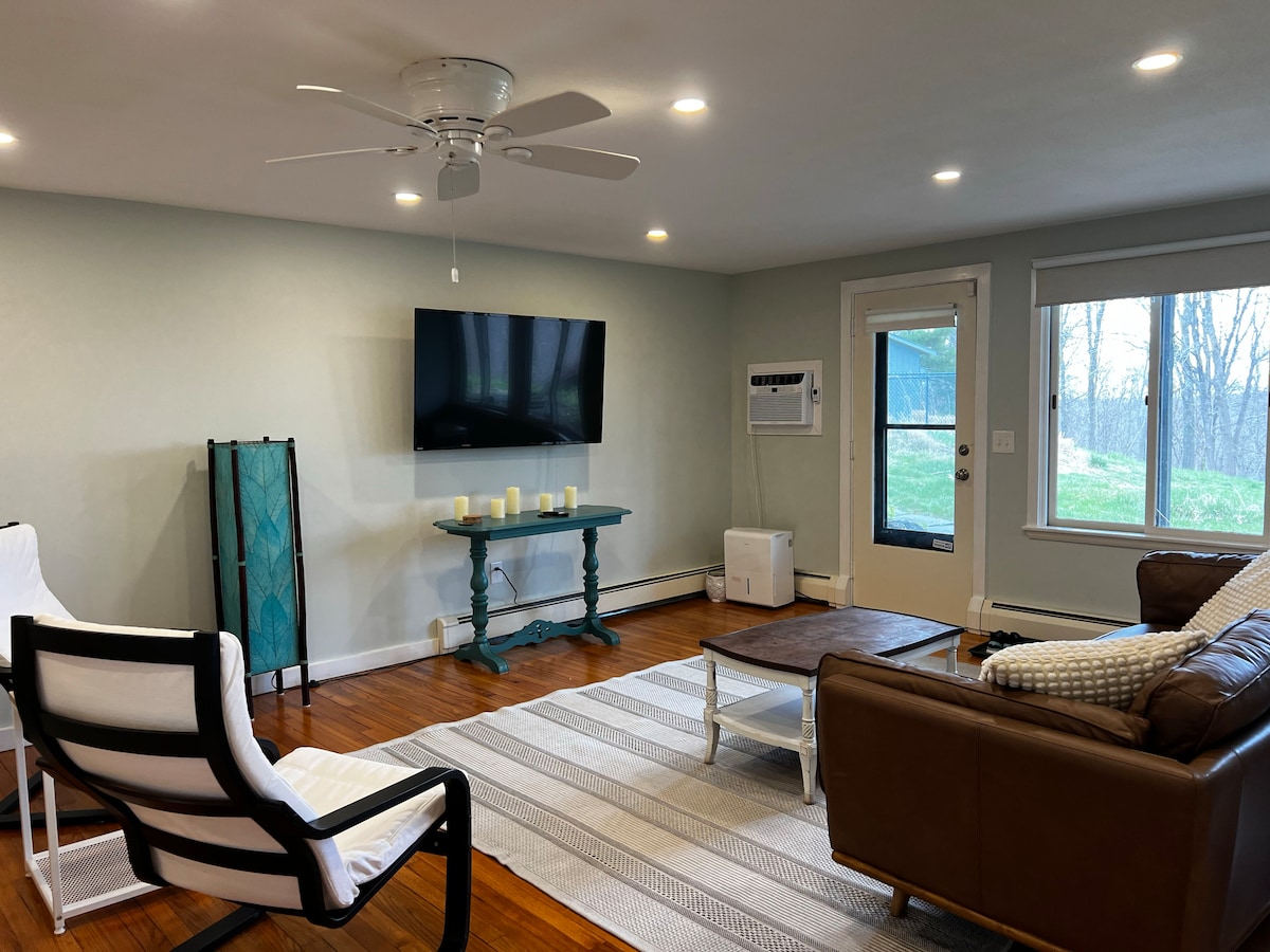 The spacious living area features comfortable seating arranged around a coffee table. A flat-screen TV is mounted on the wall, while natural light enters through the large windows. The room's neutral tones create a calming environment, complemented by a plush area rug.