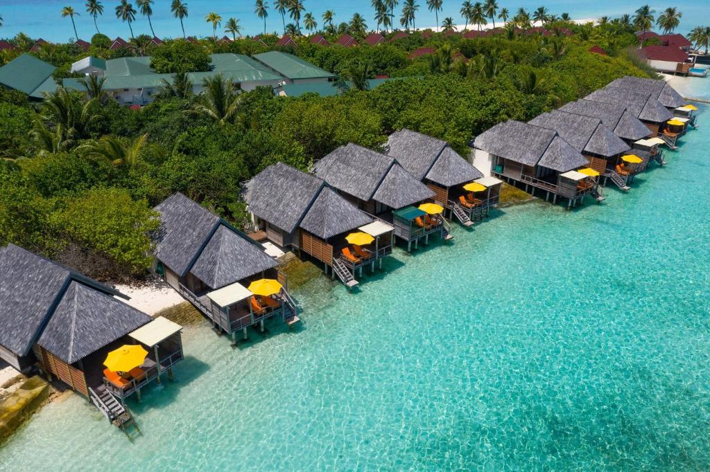 Aerial view of semi-water villas positioned along the shoreline, featuring thatched roofs and wooden decks. Each villa is complemented by an orange umbrella, offering shaded outdoor seating. Clear turquoise waters surround the villas, with lush greenery and palm trees in the background.