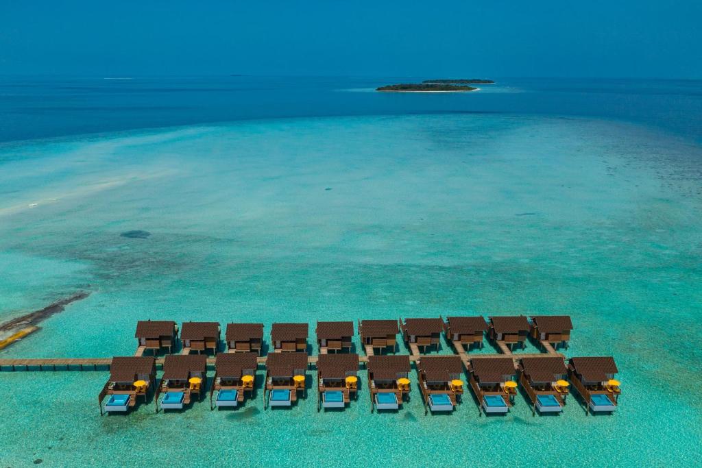 An aerial view reveals a row of water villas, each with a private pool, situated above clear turquoise waters. The vibrant ocean contrasts with a distant island, enhancing the serene coastal scenery.