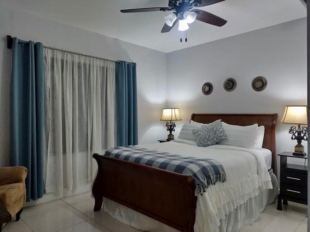 A cozy bedroom features a wooden bed with a white ruffled comforter and decorative pillows. Soft lighting is provided by two bedside lamps, and a ceiling fan enhances airflow. Light blue curtains frame a window, while three wall decorations add a touch of charm.