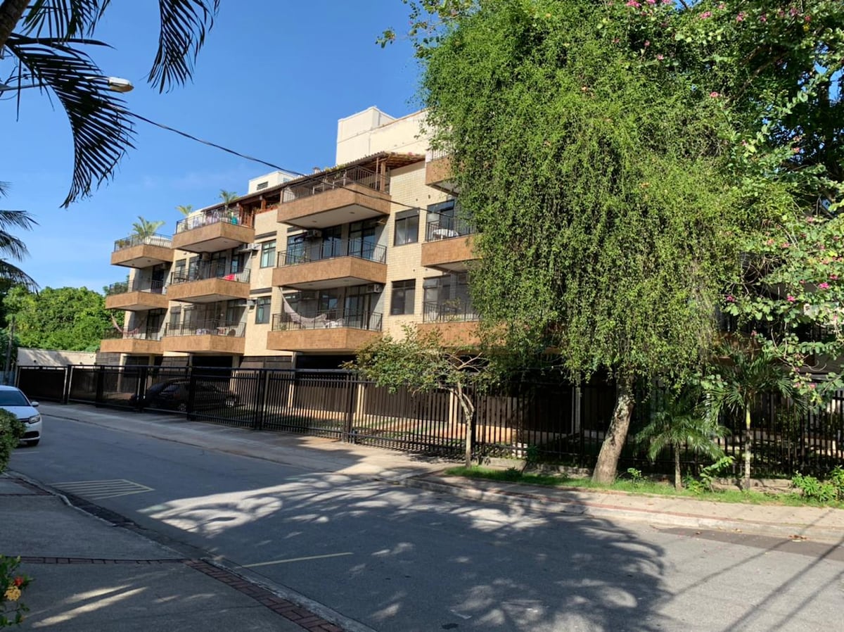The exterior of the building is captured, showcasing a three-story structure with balconies and a natural landscape of trees and shrubs. The façade reflects a blend of modern and traditional design, while a paved street runs alongside, framed by palm trees and greenery.