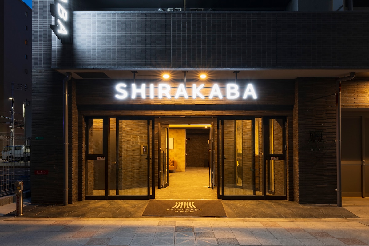 The entrance to the Shirakaba facility is visible, featuring a modern design with illuminated signage. Large glass doors allow for a clear view into the lobby area, which is softly lit, creating an inviting first impression.