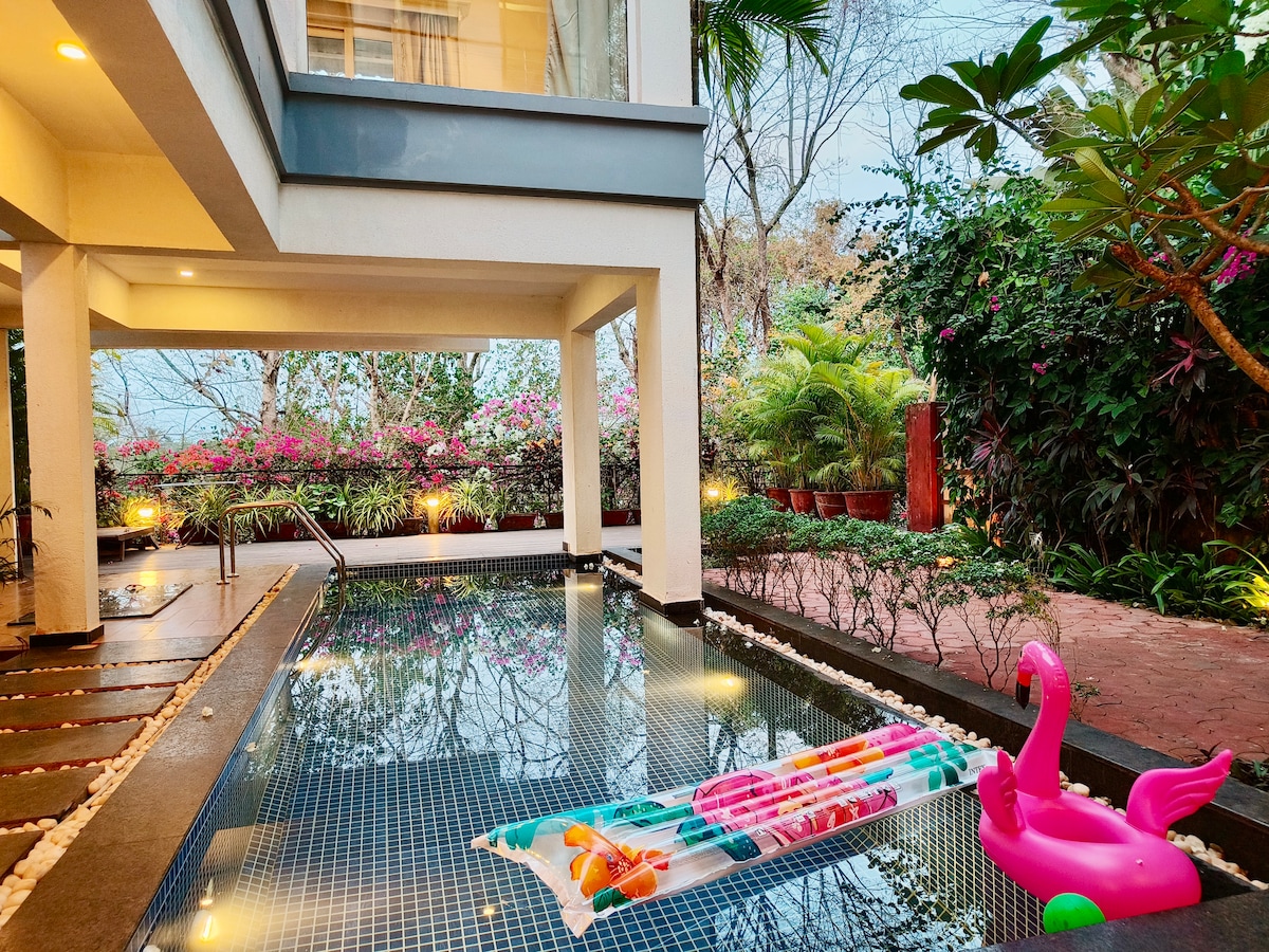 A private pool is seen alongside flower-adorned greenery, offering a serene outdoor setting. Colorful pool floats rest on the water's surface, while soft lighting highlights the area. The structure's clean lines and modern architecture blend harmoniously with the lush surroundings.