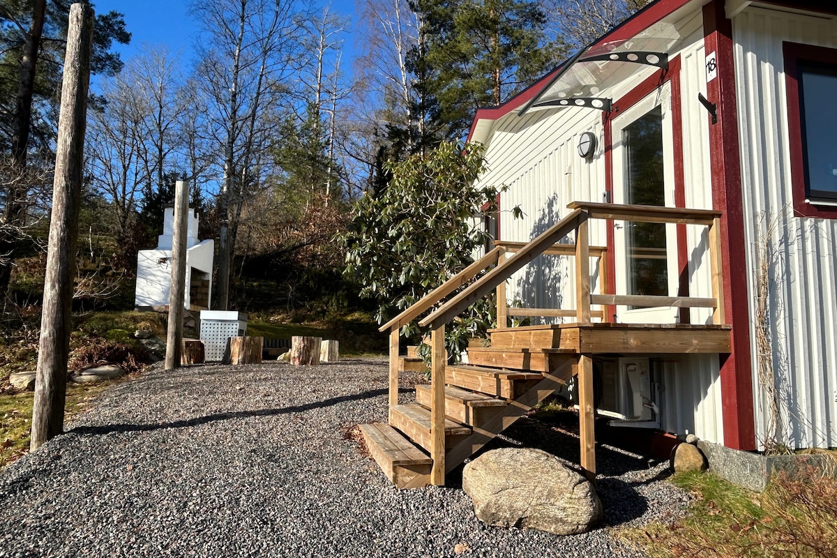 A quaint entrance is showcased with wooden steps leading to the house, surrounded by gravel and natural stone. Lush greenery is visible, and a relaxing outdoor area is highlighted in the background, complete with a wood-fired hot tub.