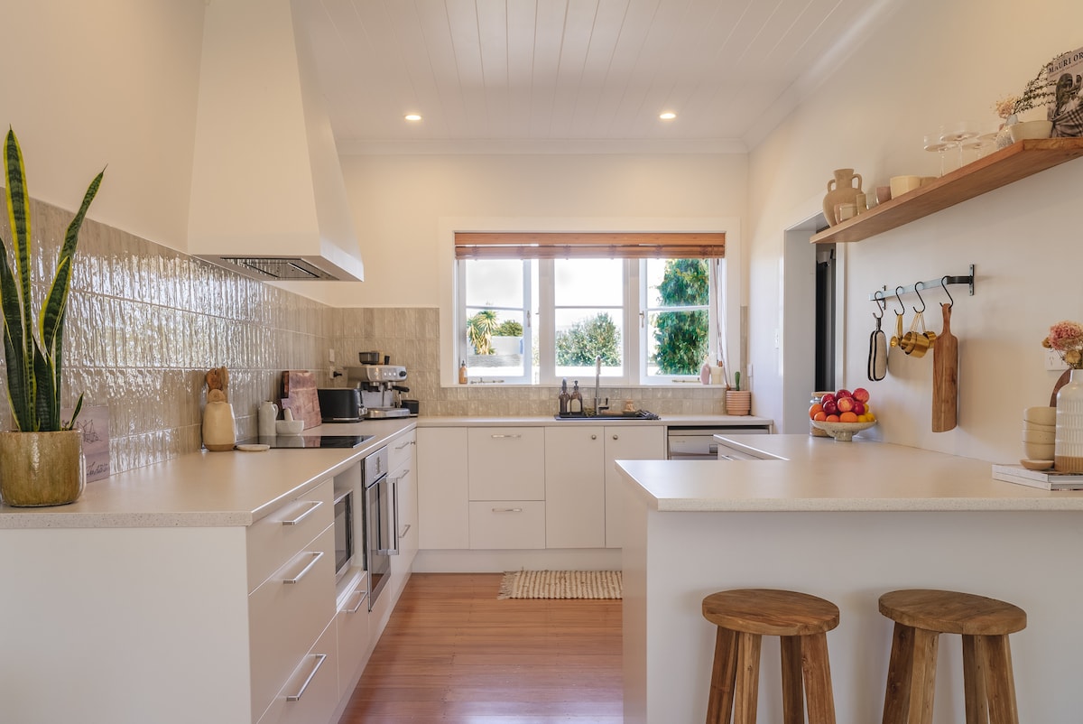 A modern kitchen features clean lines and a neutral palette. Natural light floods in through double windows, highlighting the spacious countertop and stylish bar stools. Cookware and decorative items are neatly arranged, providing both functionality and aesthetic appeal.