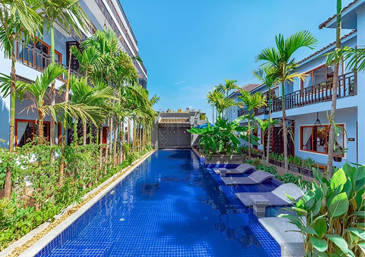 An outdoor swimming pool is surrounded by lush greenery and palm trees, creating a serene environment. Comfortable lounge chairs are positioned along the edge of the pool, while vibrant plants contribute to the tranquil setting under clear blue skies.