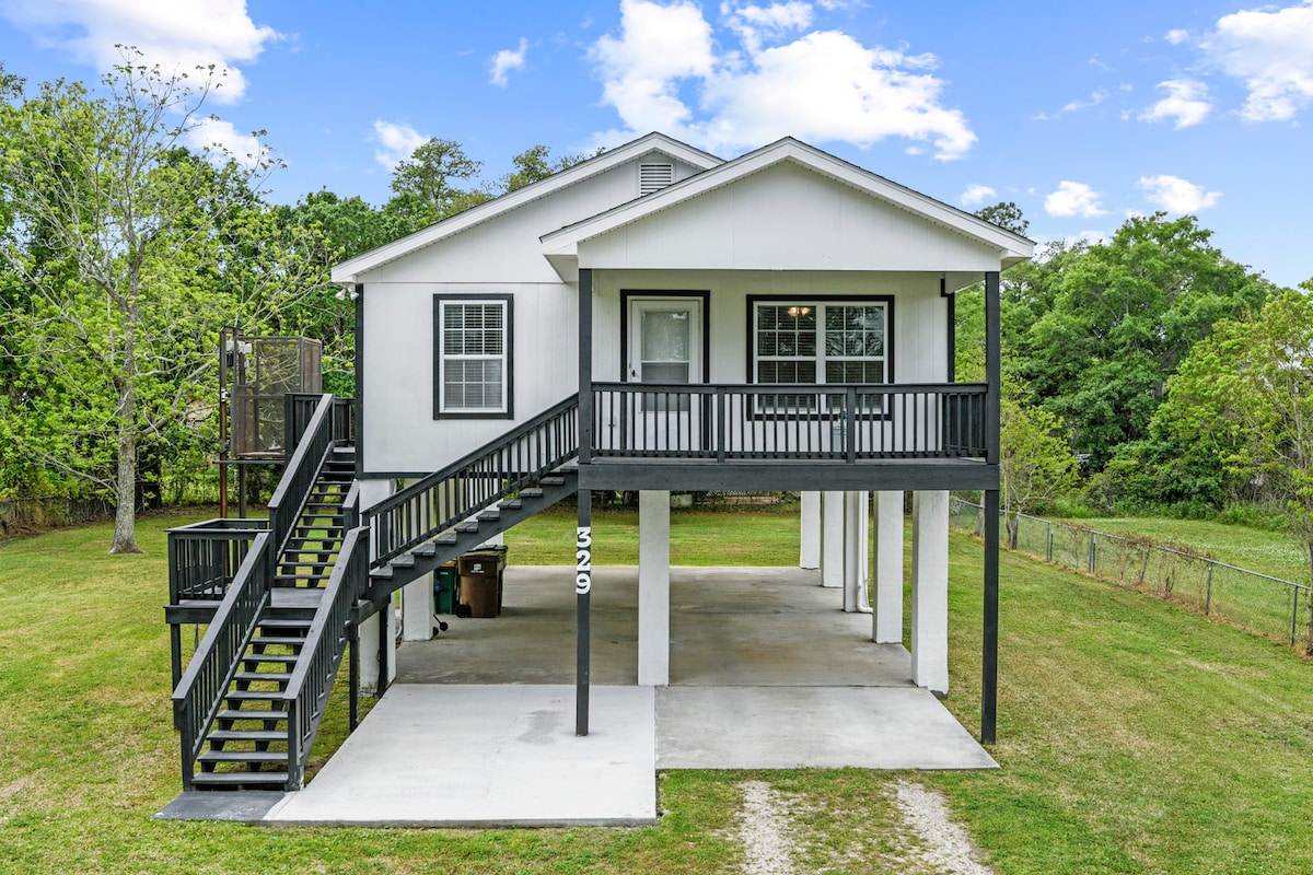 A two-story house is presented on stilts, featuring a deck with black railings and stairs leading to the entrance. The property is surrounded by a grassy yard and trees, providing a natural setting. A paved area is visible beneath the house.