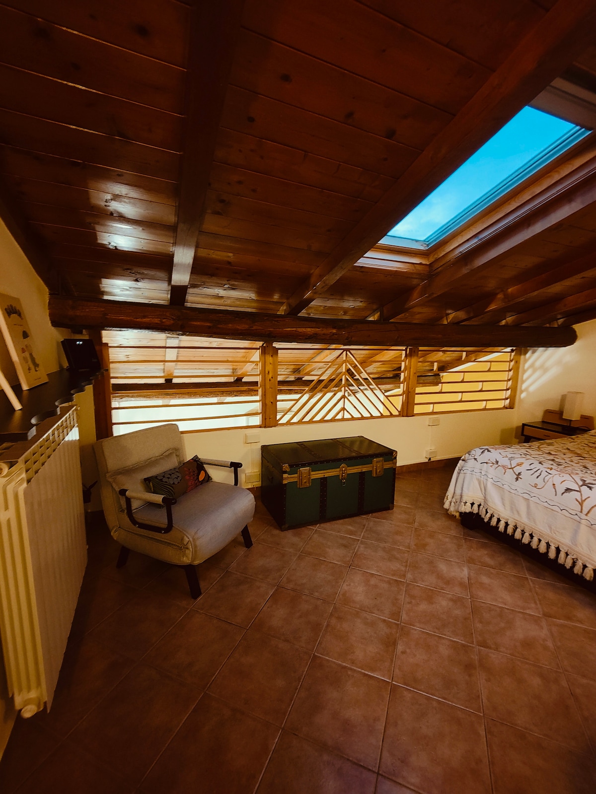 A spacious room features wooden ceiling beams and a skylight that allows natural light to fill the space. A cozy chair is positioned near a decorative trunk, enhancing the room's functionality. The floor is tiled, contributing to a warm and welcoming ambiance.