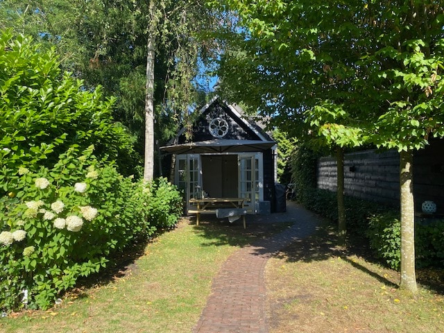 A charming garden cottage is framed by lush greenery, featuring a welcoming path made of brick leading to its entrance. Large windows allow natural light to fill the interior, and a spacious outdoor area includes a wooden table surrounded by trees.