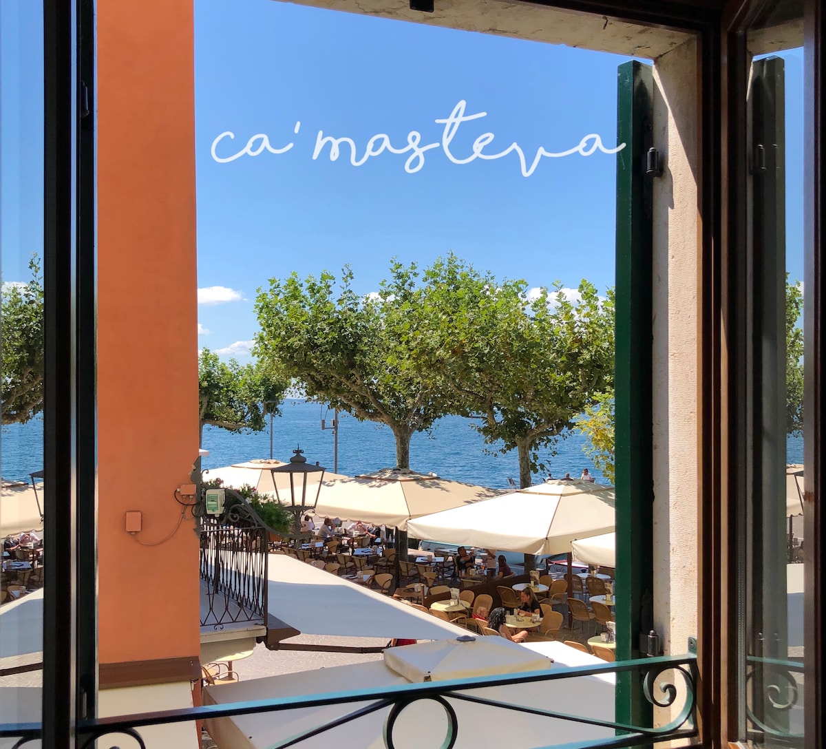 A view from the window showcases the vibrant waterfront with outdoor seating beneath large umbrellas. Lush trees line the shore, and glimpses of the lake are visible, enhancing the atmosphere of the bustling area below.