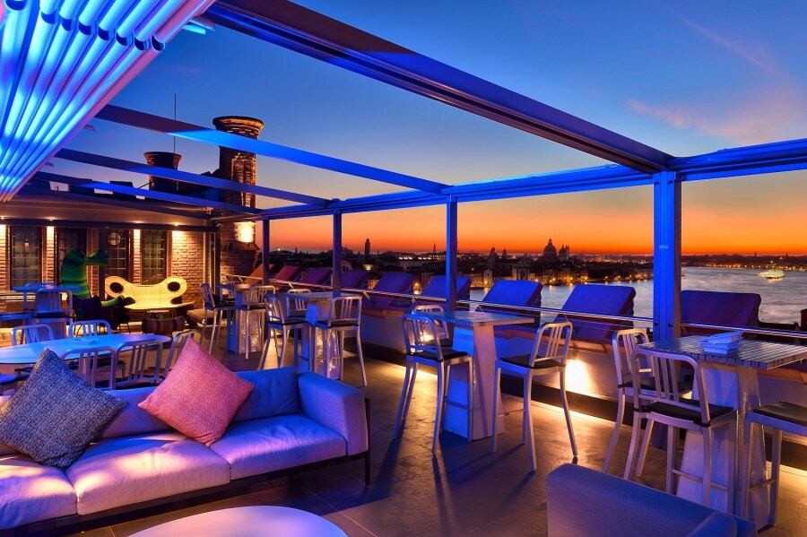 A rooftop lounge area is illuminated by colorful LED lights as the sun sets over the skyline. Plush sofas are arranged around low tables, with vibrant cushions adding a cozy touch. The panoramic views of the city are visible against a backdrop of a twilight sky.