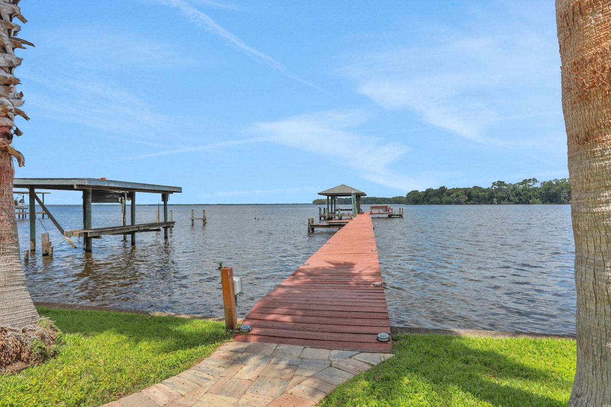 A wooden dock extends over the calm St. Johns River, leading to a covered gazebo. The shoreline is bordered by lush green grass, with several palm trees providing shade in the foreground. The clear blue sky complements the tranquil water.