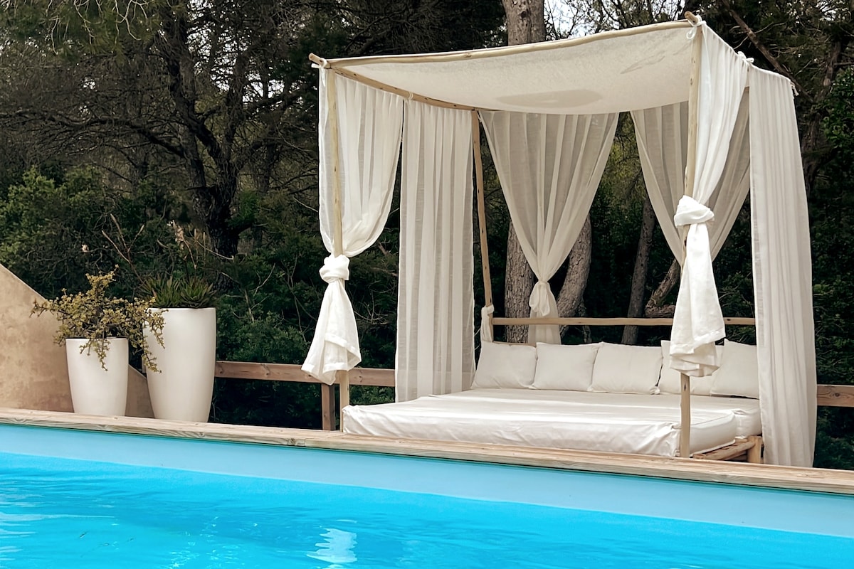 A sheltered lounging area beside a sparkling pool features a wooden frame draped in white fabric. Plush cushions are arranged for comfort, and large planters with greenery stand nearby, enhancing the serene outdoor setting.