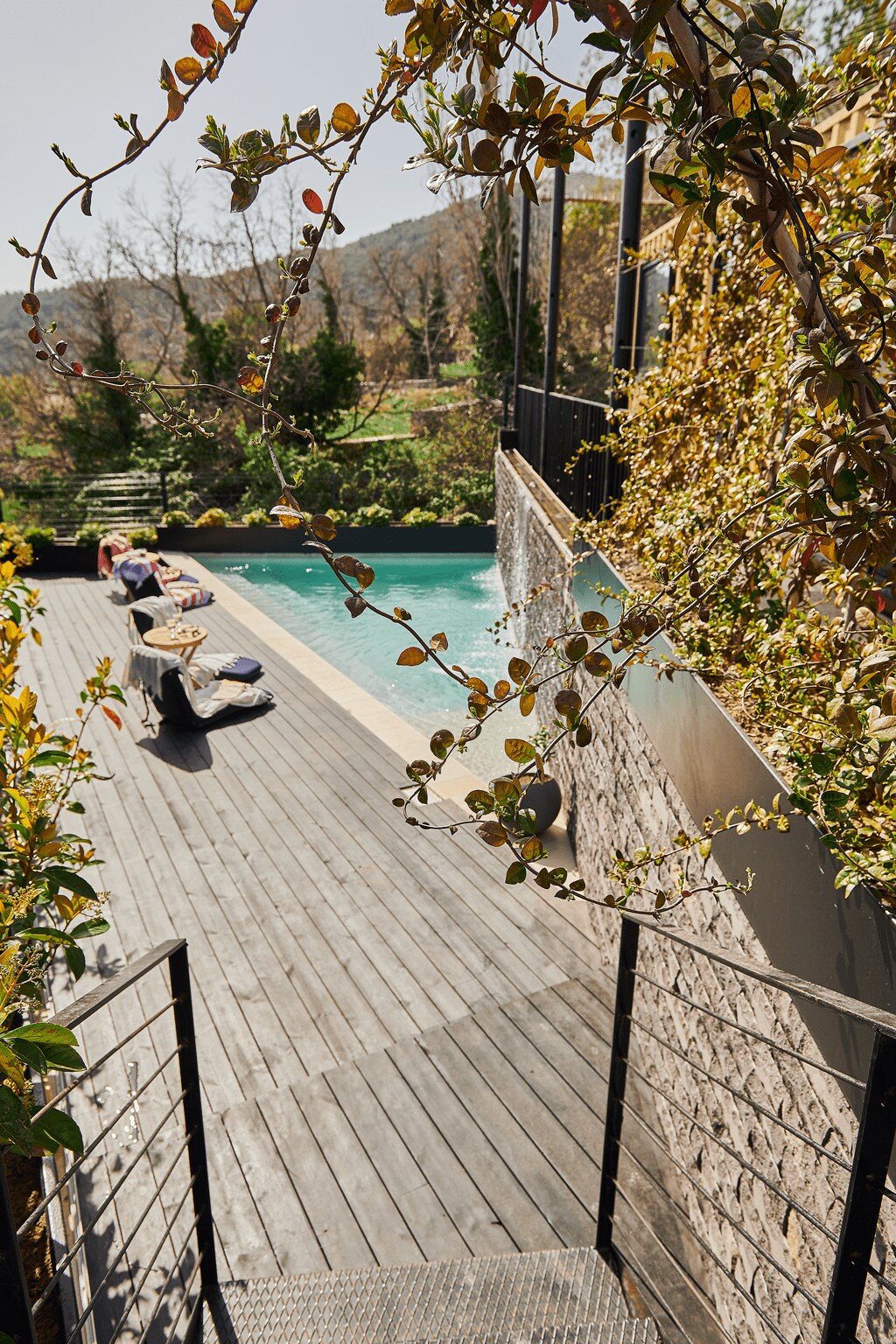A serene outdoor area features a polished wooden deck that leads to a shimmering pool. Lush greenery surrounds the space, and lounge chairs are positioned nearby for relaxation. Steps descend from the deck, blending harmoniously with the natural landscape.