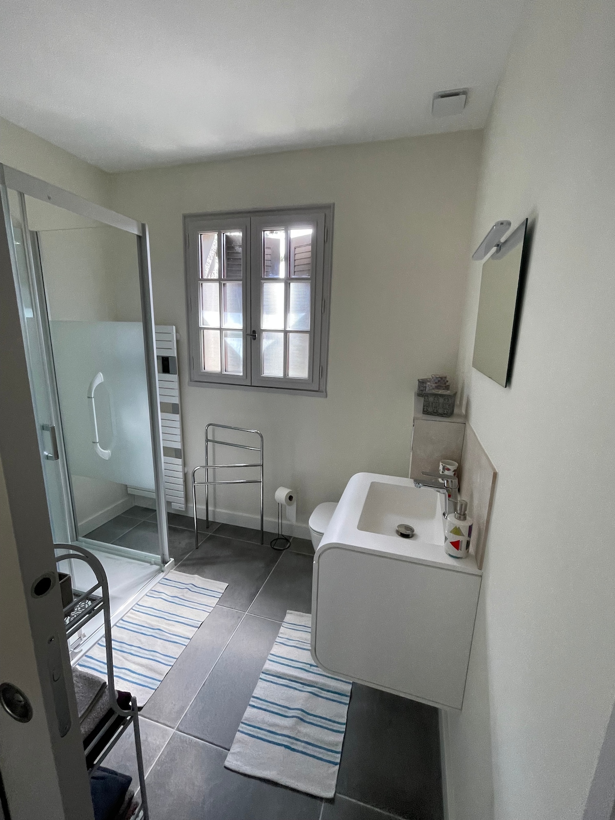 A modern bathroom is presented, featuring a walk-in shower with a glass panel and a contemporary sink unit. Two striped bath mats are placed on the tiled floor. A window with multiple panes allows natural light, while a towel warmer and a mirror add functionality.