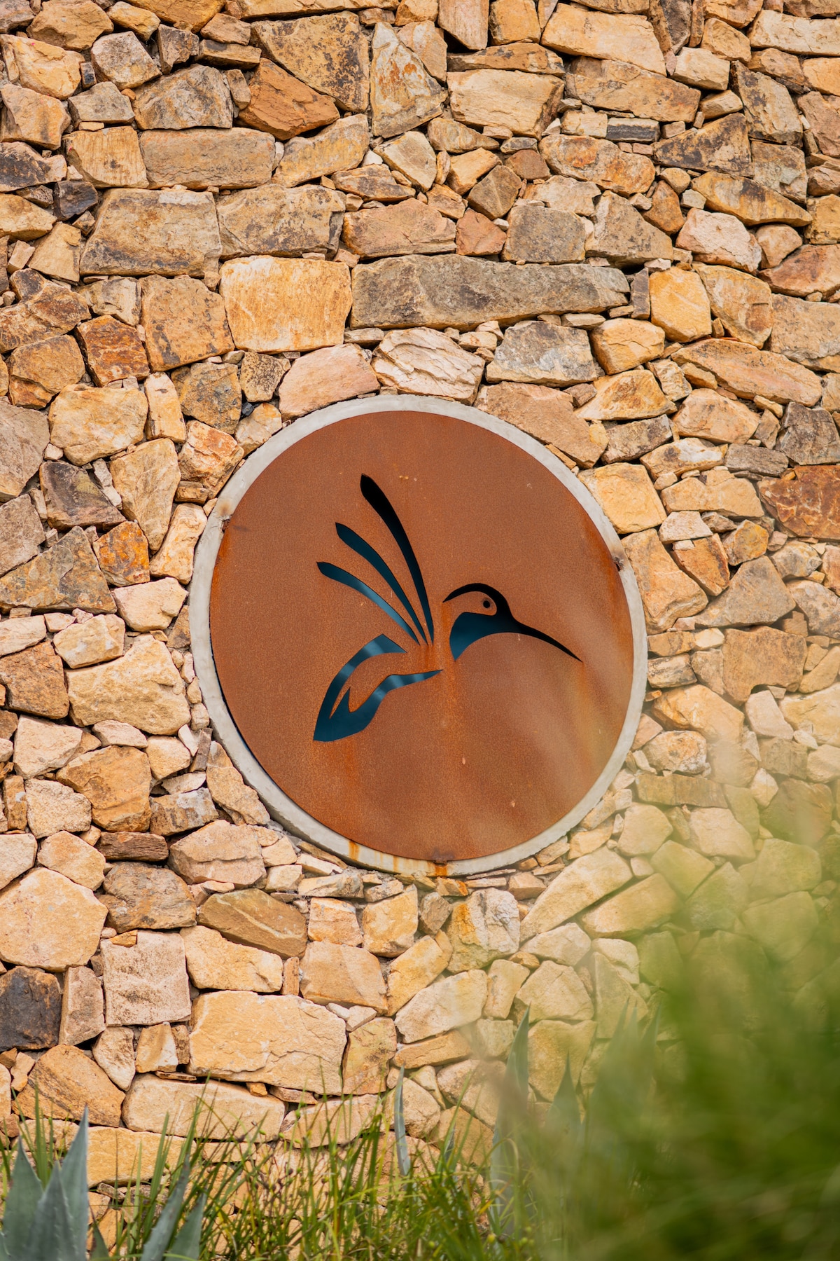 An artistic metal emblem of a hummingbird is mounted on a stone wall. The circular design contrasts with the textured surface of the rustic stones, which vary in shape and color, enhancing the natural surroundings.