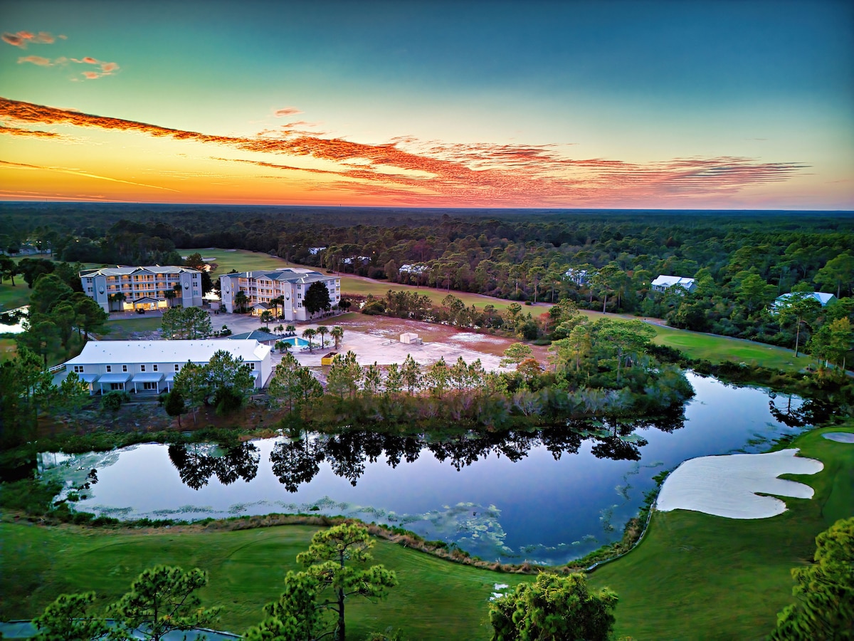 A sweeping view captures the serene landscape at sunset, featuring a tranquil pond reflecting the vibrant sky. Multiple buildings are visible, surrounded by lush greenery, while the expansive horizon reveals a blend of forest and open spaces, creating a peaceful atmosphere.