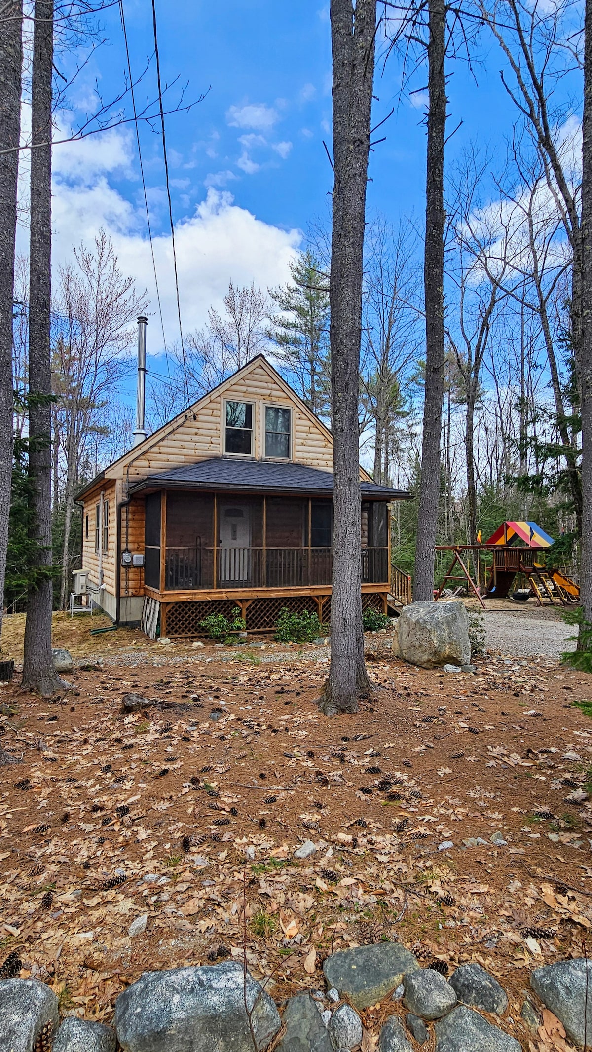 A log cabin is presented amid a wooded area, with a screened porch accessible via a wooden step. Play equipment is visible nearby, framed by tall trees and natural ground cover. The sky is partly cloudy, allowing sunlight to highlight the cabin's exterior.