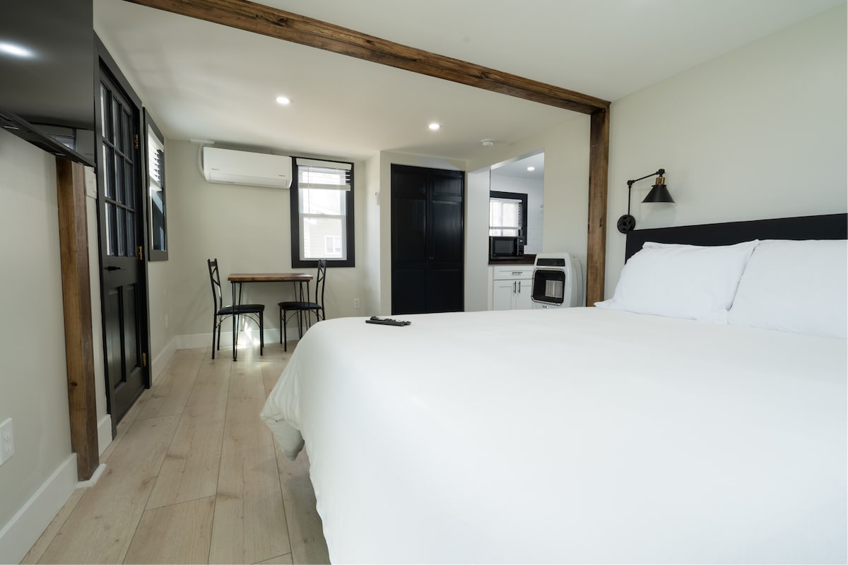 The interior of the cabin is shown, featuring a cozy sleeping area with a spacious bed dressed in white linens. A small dining table for two and a kitchenette are visible in the background, along with natural light illuminating the sleek, modern design.