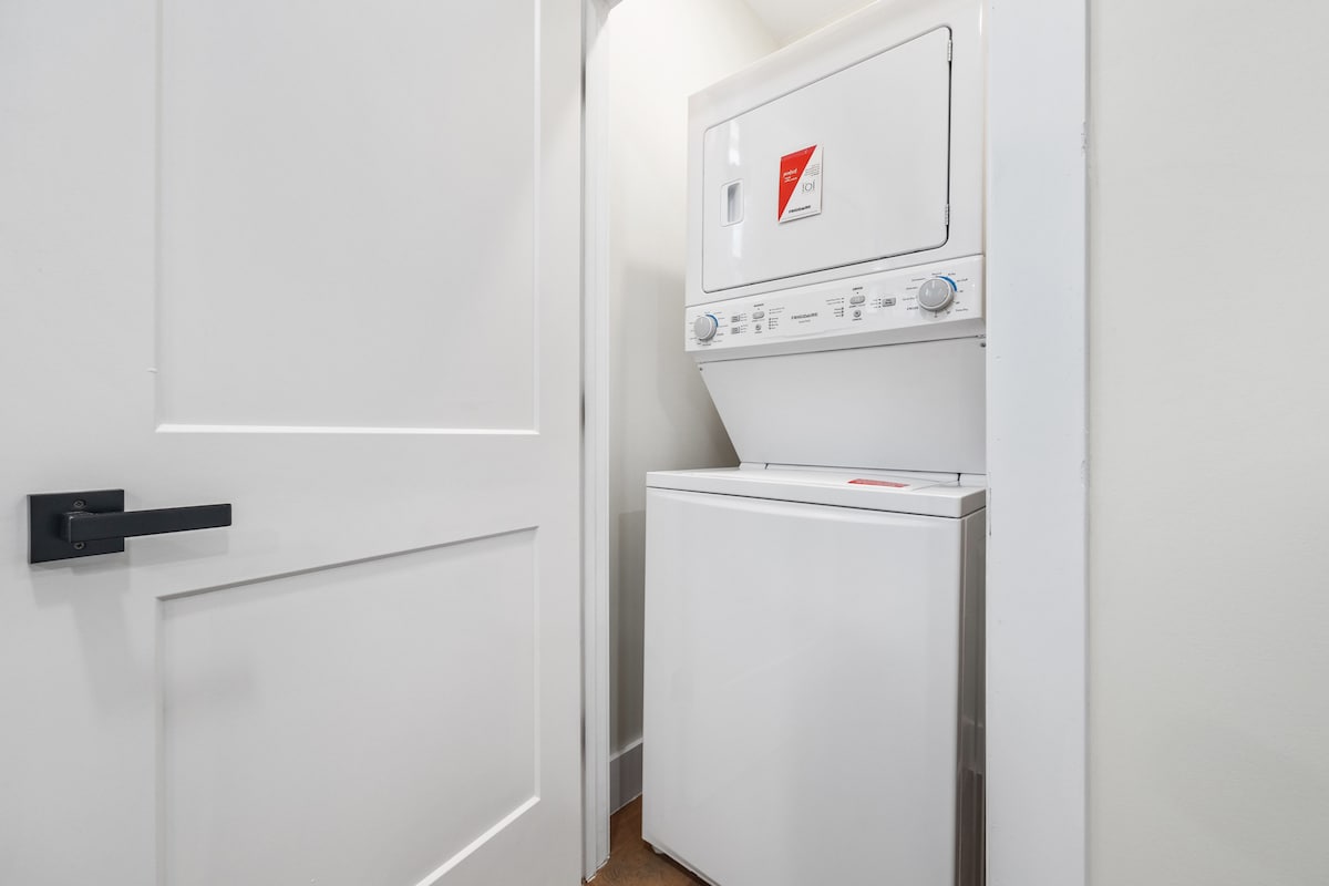 A compact laundry space is presented, featuring a stacked washer and dryer unit. The appliances are situated against a clean, light-colored wall, with a white door partially open, allowing access to the laundry area.
