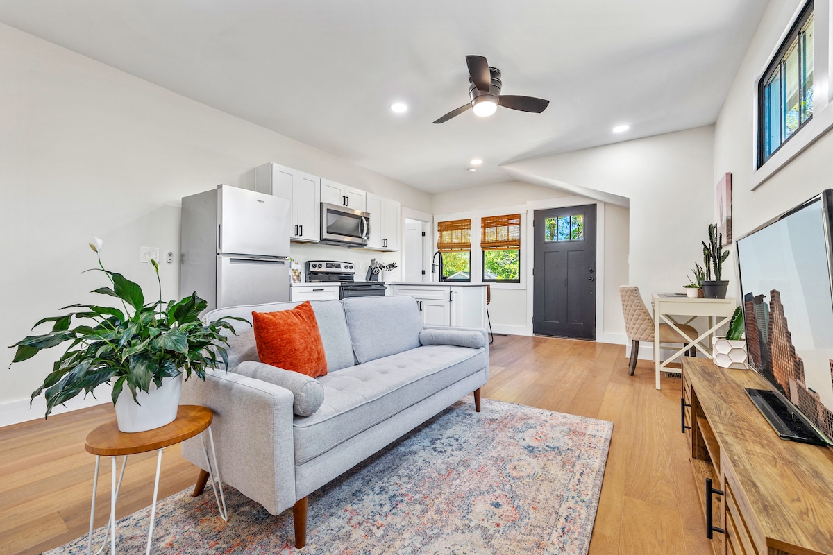 A modern living area features a light grey sofa with an orange accent pillow, complemented by a patterned area rug. A wooden coffee table stands nearby, with a potted plant adding greenery. The kitchen showcases white cabinets and stainless-steel appliances, illuminated by natural light.