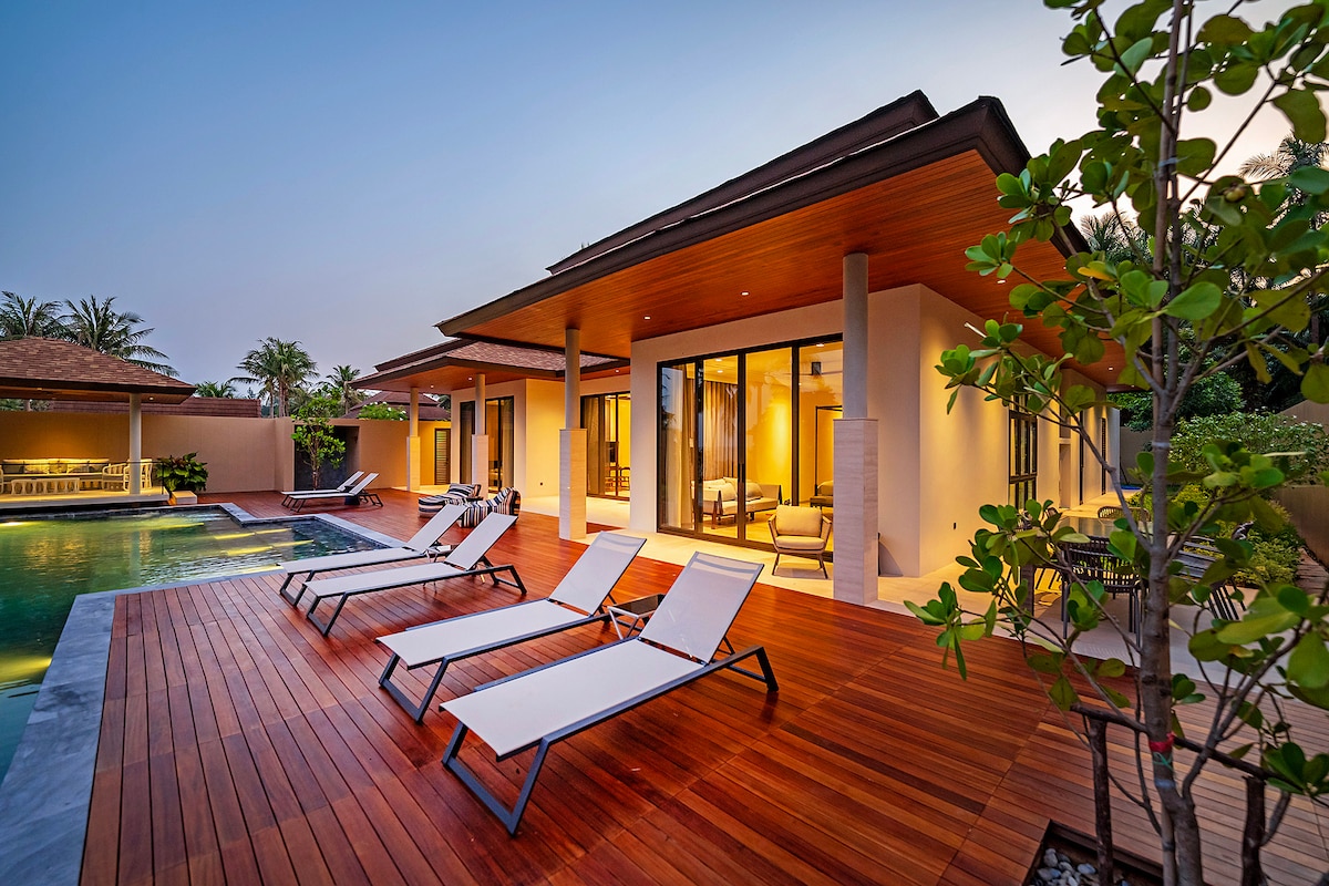 A wooden deck surrounds a private infinity pool, with four lounge chairs arranged for relaxation. The villa's expansive glass doors offer a view into a spacious living area. Lush greenery is visible along the perimeter, providing a tropical backdrop to the setting.