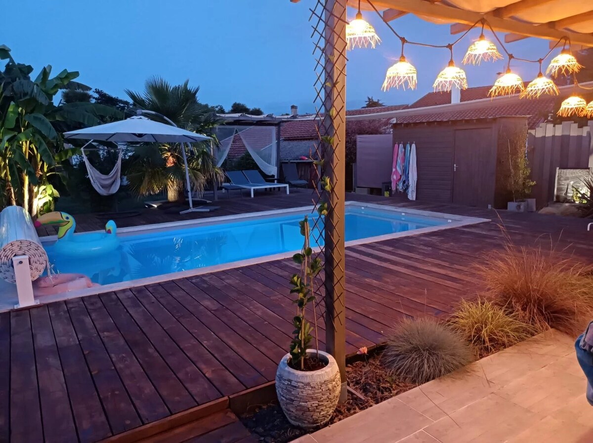 The heated swimming pool, illuminated gently at dusk, features a surrounding wooden deck. Comfortable lounge chairs are positioned beside the water, with umbrellas offering shade. Decorative lights hang from the pergola, enhancing the inviting outdoor space. Potted plants add a touch of greenery nearby.