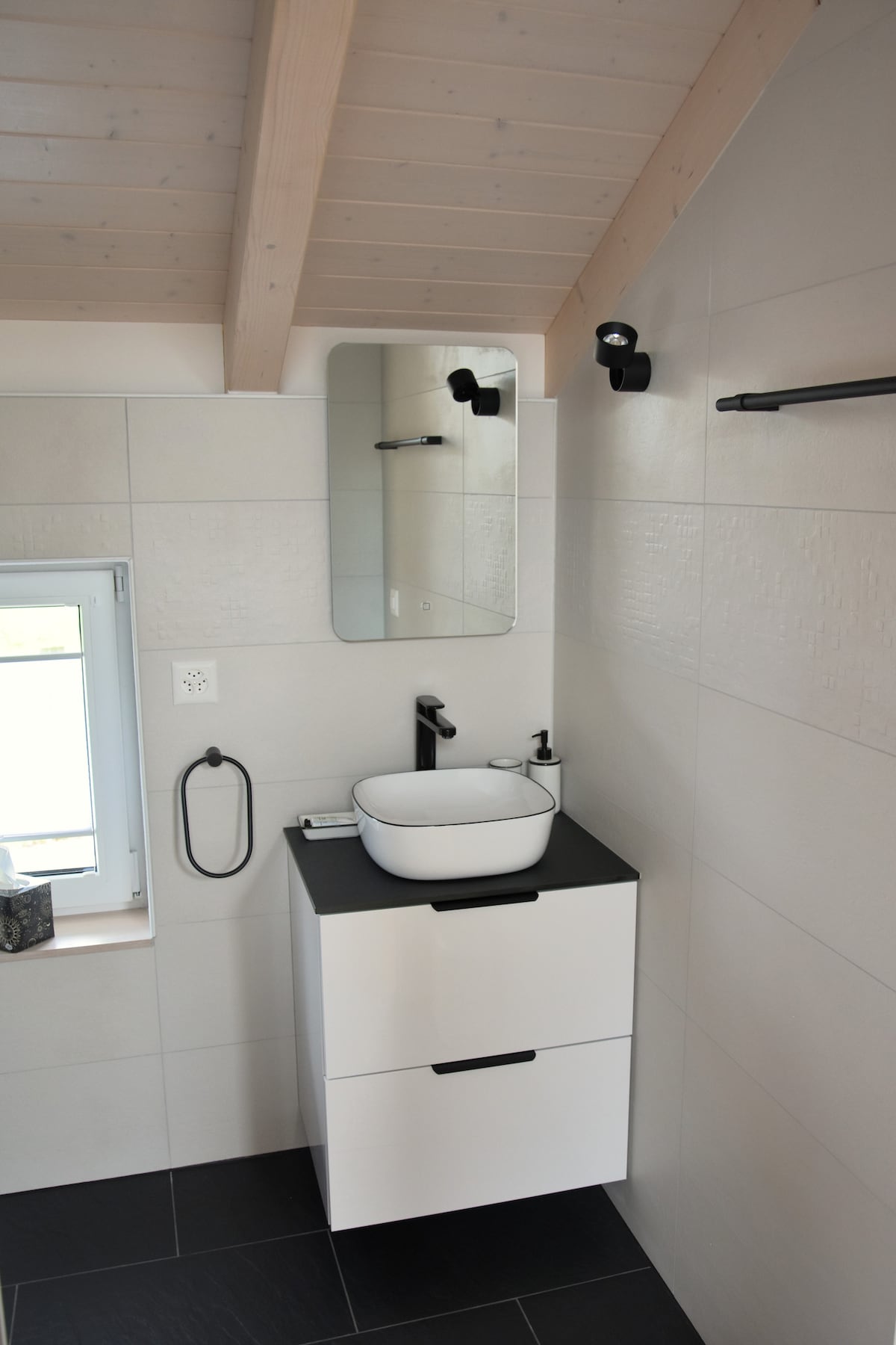 A modern bathroom is presented with a white wall-mounted vanity featuring a black countertop and a round basin. A large mirror above reflects the space. A window allows natural light to enter, complemented by minimalist black fixtures and a towel holder.