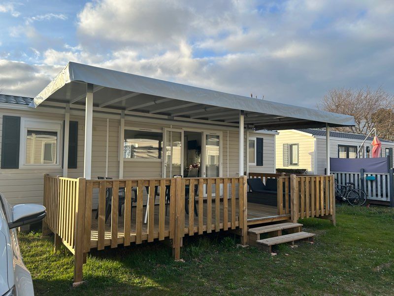 A spacious mobile home is presented with a covered terrace surrounded by a wooden railing. Outdoor seating is available on the terrace, inviting relaxation. The entrance is highlighted by a few steps that lead to a light-colored façade, complemented by green shutters.